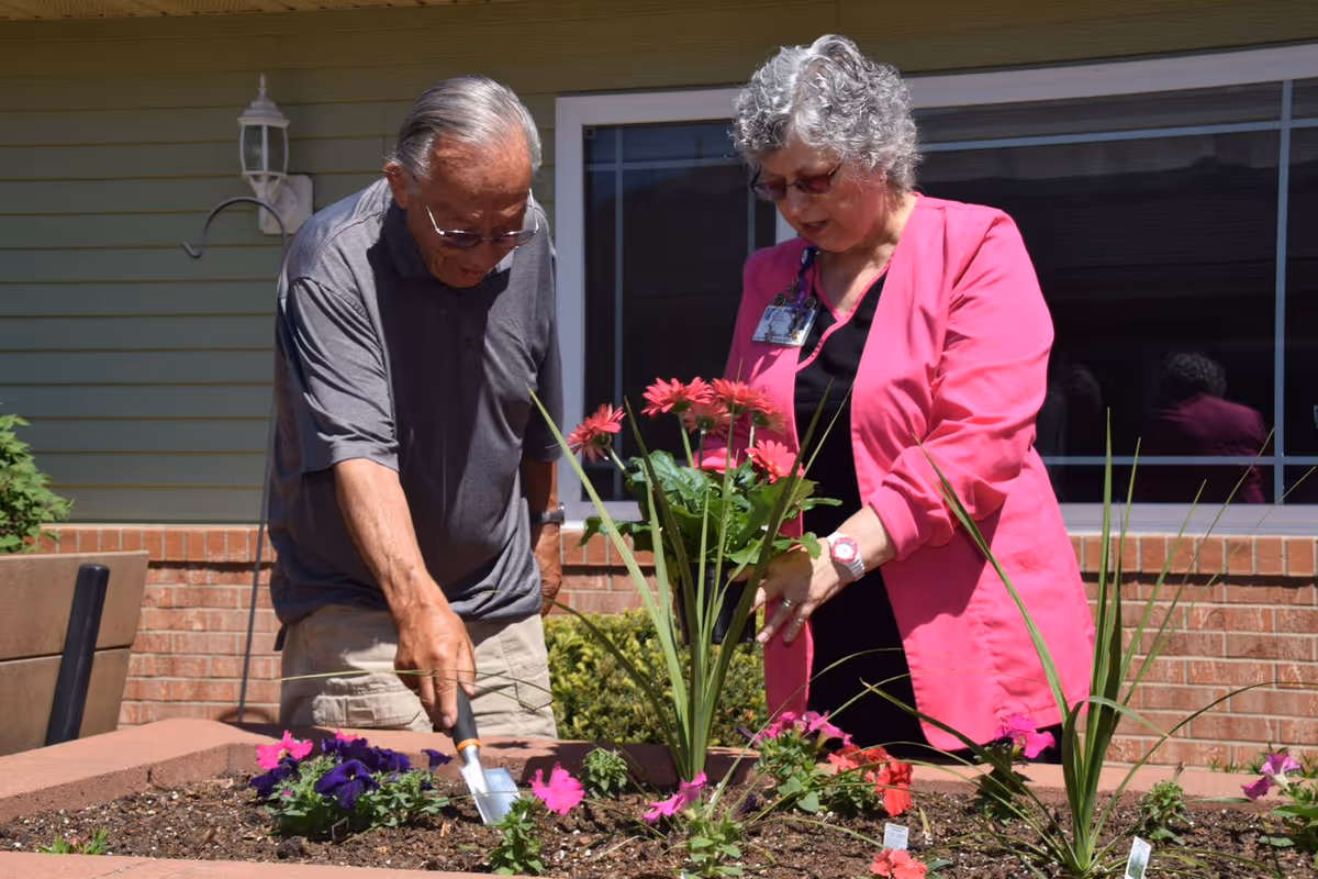 An elderly man and a woman with gray hair wearing a bright pink jacket are gardening together in a raised flower bed outside a building with a brick wall and window in the background. The man is using a small gardening trowel to tend the soil while the woman looks on and holds some flowers.