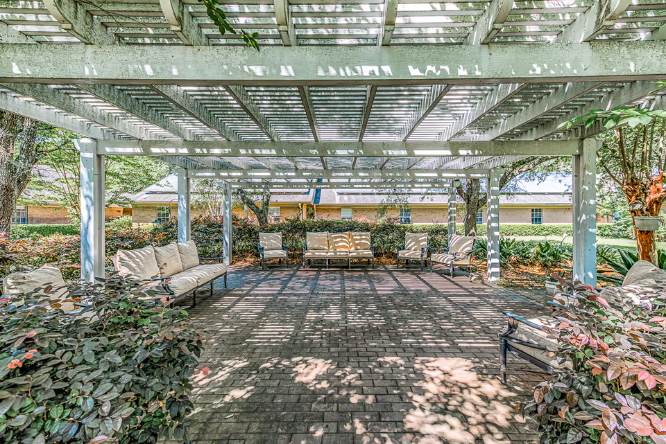 Shaded outdoor pergola with cushioned benches and chairs on a brick patio surrounded by landscaping.