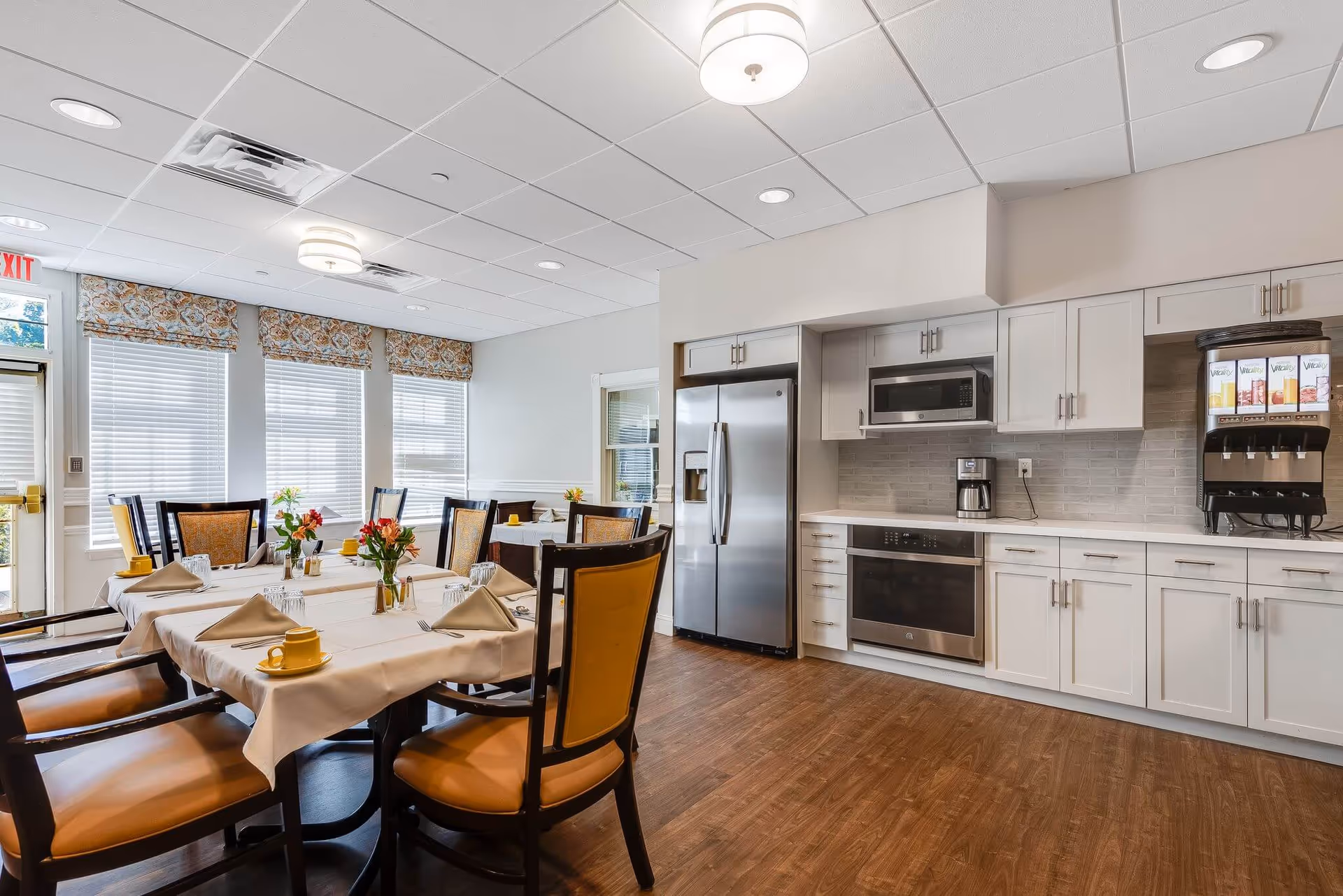 Well-lit communal dining area with set tables and chairs adjacent to a modern kitchen with stainless steel appliances.