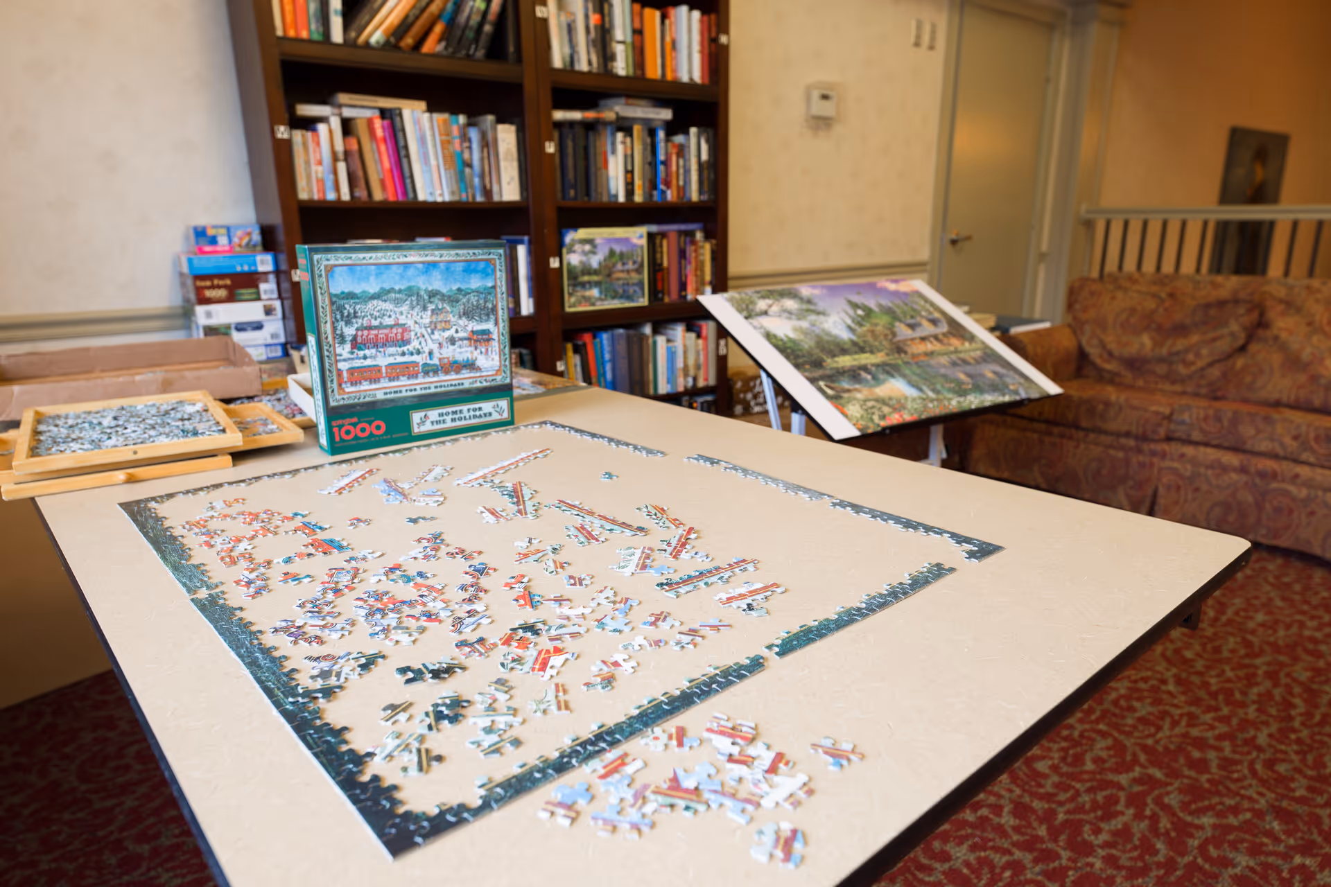 A table with an incomplete jigsaw puzzle in progress, surrounded by puzzle pieces and puzzle trays. Behind the table is a bookshelf filled with books and board games, a couch with patterned upholstery, and a door in the background. The room has a carpet with a red and gold pattern.