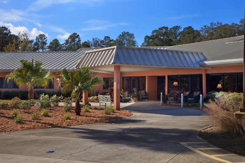 Exterior view of Wellsprings Residence LLC showing the entrance area with a covered driveway, landscaped garden with palm trees and shrubs, and large windows revealing a seating area inside.