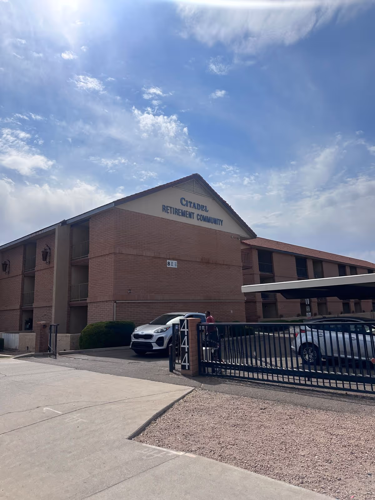Three-story brick building labeled "Citadel Retirement Community" with gated parking and cars under a partly cloudy sky.