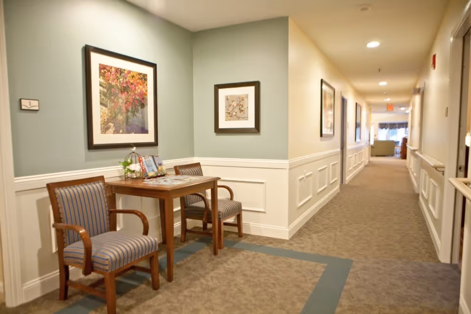 A well-lit hallway in a senior living facility with beige and green walls, carpeted floor, and white wainscoting. On the left side, there is a small wooden table with two striped upholstered chairs. Above the table are two framed floral artworks. The hallway extends into the distance with more framed pictures on the walls and a seating area visible at the far end.