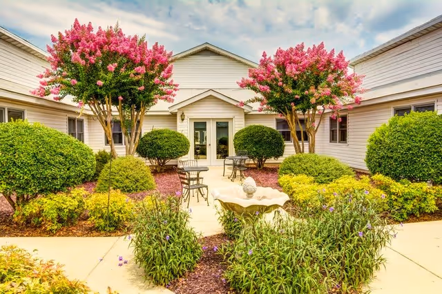 Outdoor courtyard area at Carolina Gardens at West Columbia featuring a paved walkway, green bushes, flowering pink trees, and metal tables with chairs. The courtyard is surrounded by light-colored building walls with windows and a central door.
