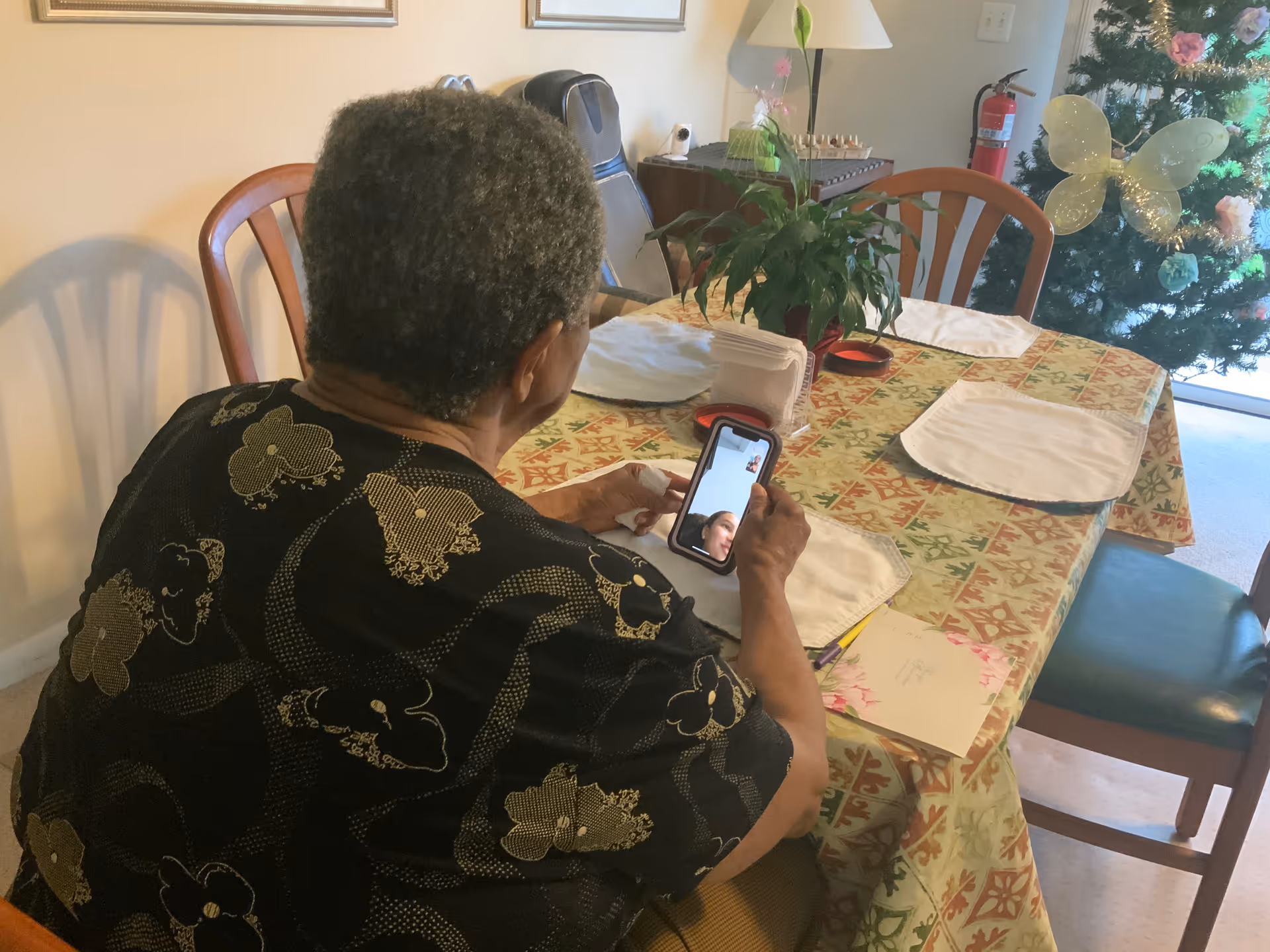 An elderly person sitting at a dining table with a patterned tablecloth, holding a smartphone and video chatting with another person. The table has placemats, a plant, and some papers. In the background, there is a decorated Christmas tree and a fire extinguisher on the wall.