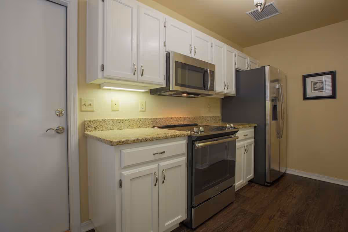A kitchen with white cabinets, granite countertops, a stainless steel microwave, stove, and refrigerator. The walls are painted beige, and there is a framed picture hanging on the wall. The floor is wooden, and there is a white door on the left side of the image.