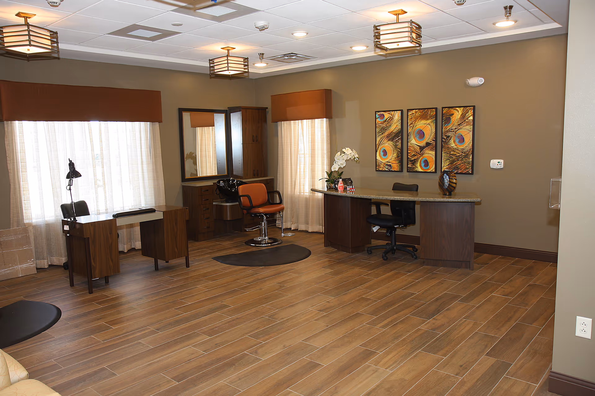 A spacious interior room with wood-look tile flooring and beige walls. The room features a reception desk with a black office chair, a salon chair with a mirror and cabinet behind it, and a small desk with a lamp and chair near large windows covered with sheer curtains and brown valances. Three colorful peacock feather artworks hang on the wall above the reception desk. The ceiling has recessed lighting and decorative light fixtures.