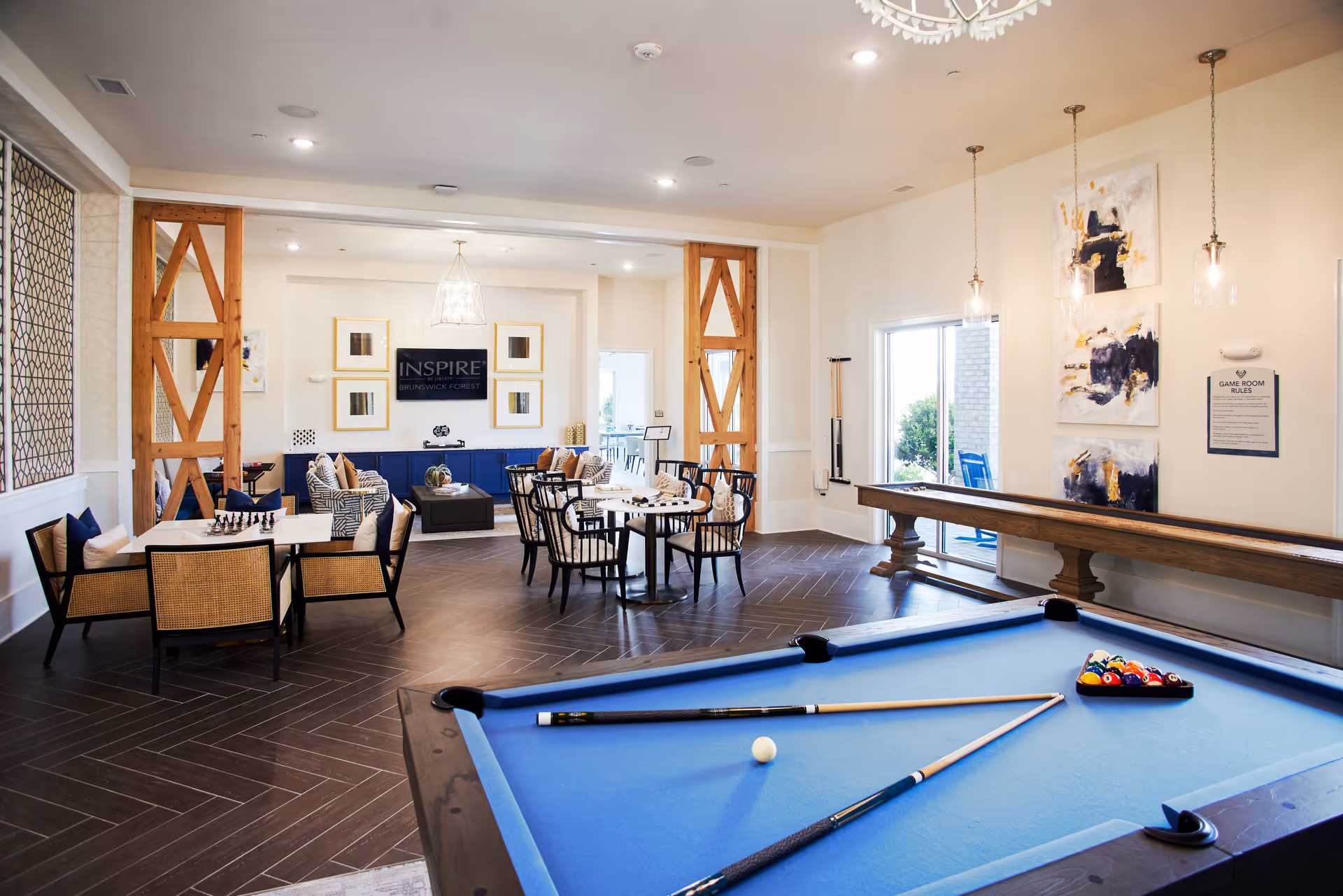 Community game room with a blue pool table in the foreground, shuffleboard and seating areas, and an 'Inspire Brunswick Forest' sign on the far wall.