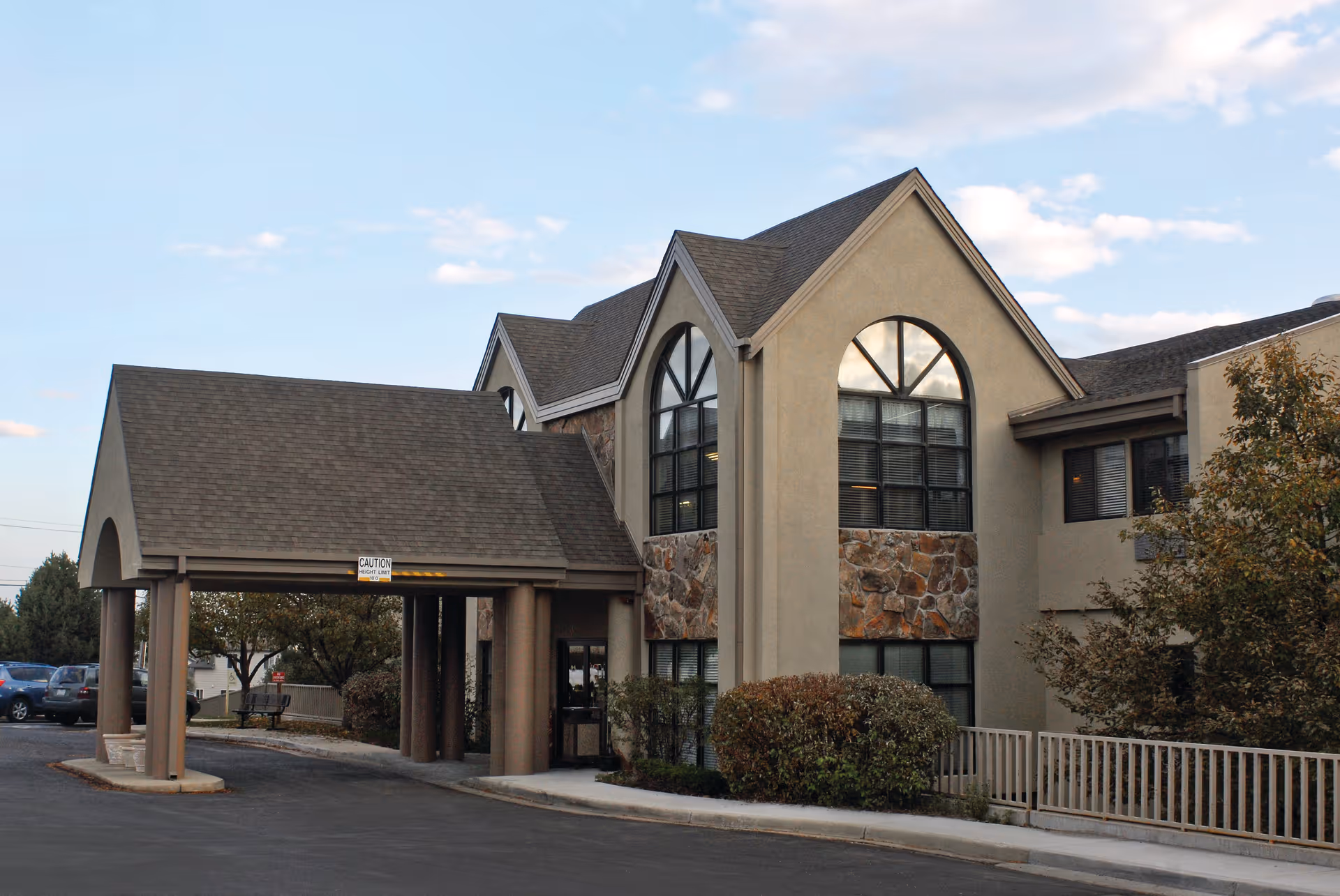 Front exterior of a two-story senior living facility with a covered drop-off, arched windows, and stone accents.