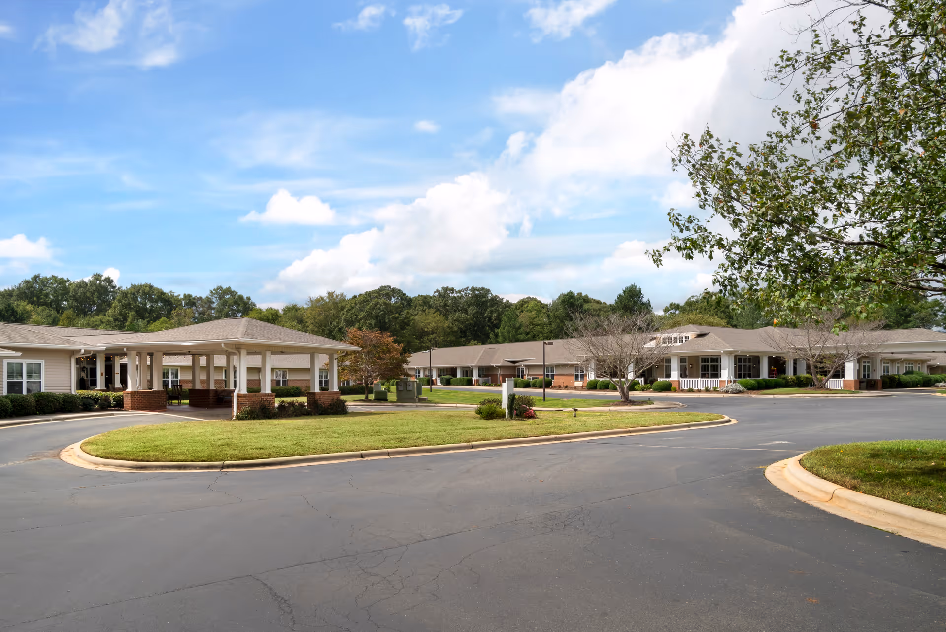 Front exterior of a single-story senior living facility with a circular driveway, covered entrance canopy, and landscaped grounds under a blue sky.
