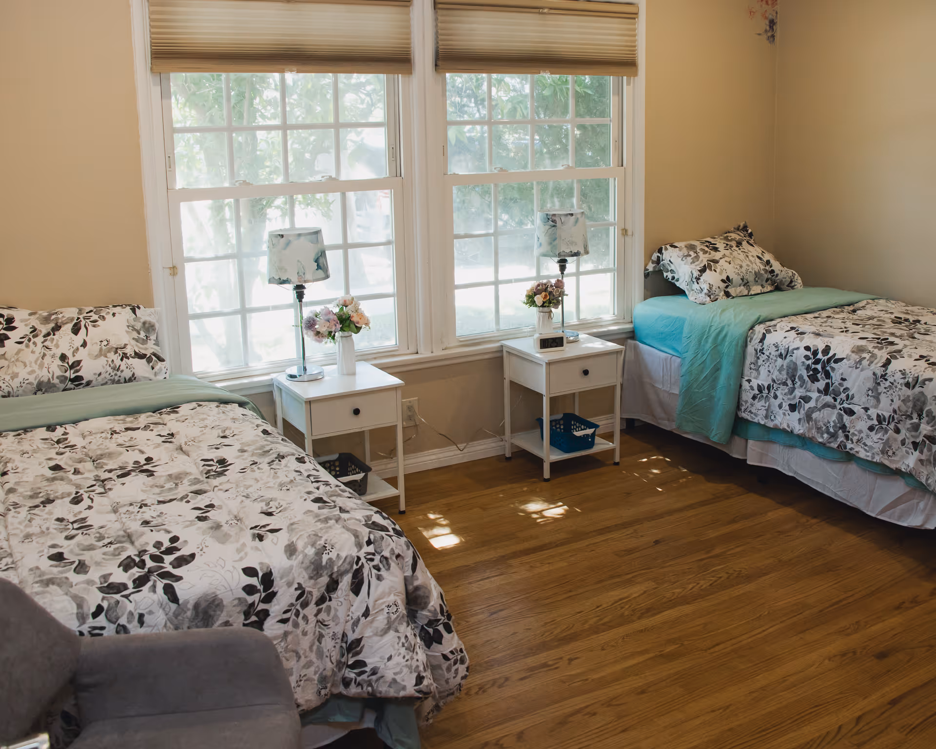 A bright bedroom with two twin beds featuring floral bedding, bedside tables with lamps and flowers, and large windows.