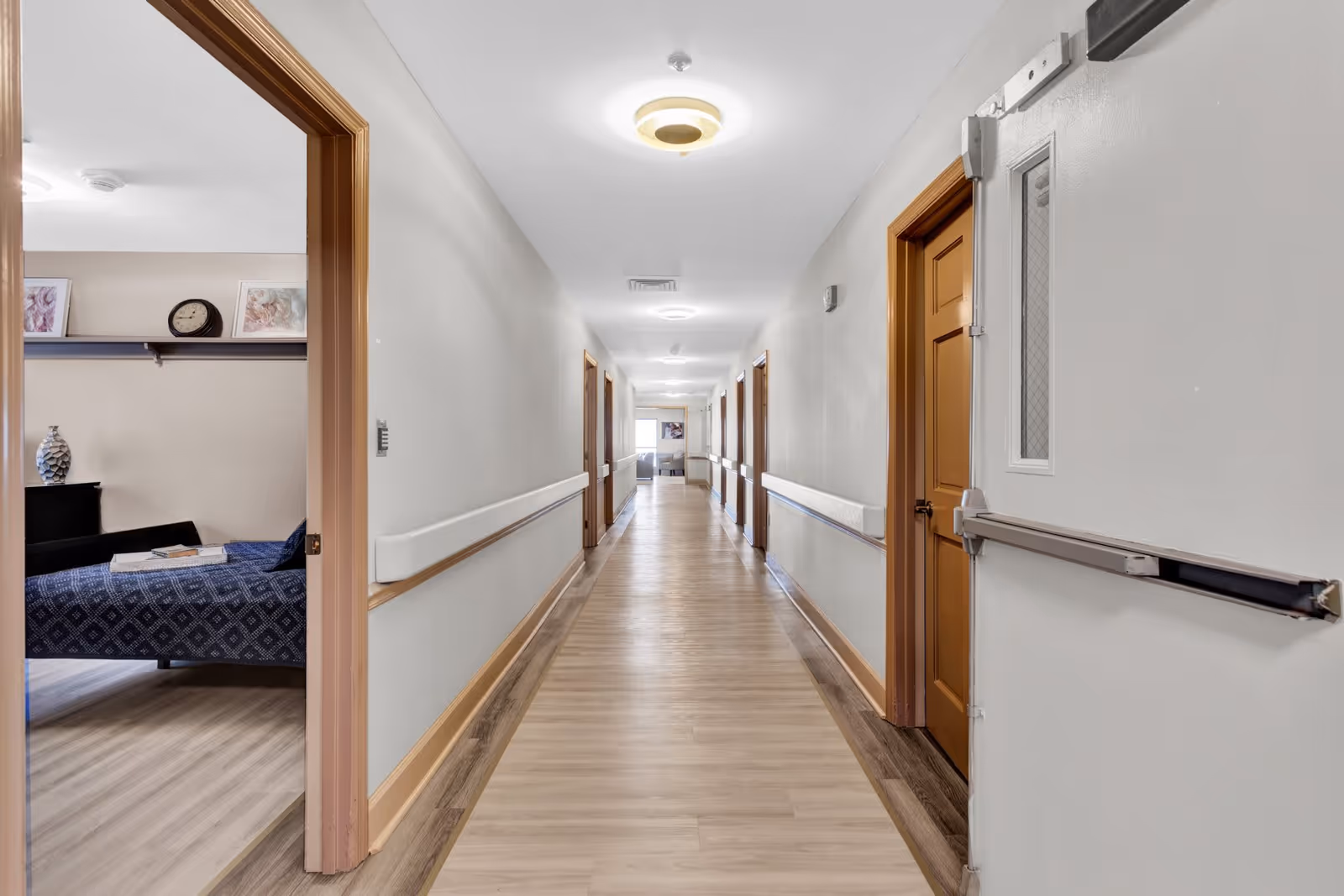A long, well-lit hallway in an assisted living facility with light-colored walls and wood flooring. Several closed wooden doors line the right side of the hallway, while the left side has an open door revealing a bedroom with a bed covered in a blue patterned blanket, a clock, and framed pictures on a shelf. Handrails run along both sides of the hallway.