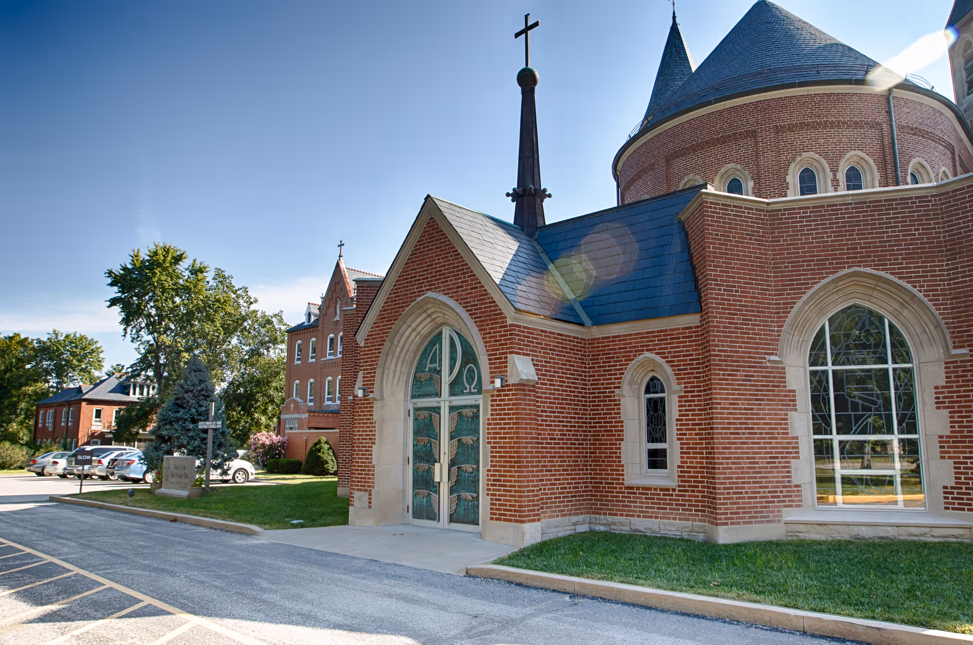 Exterior view of a red-brick church-like building with arched windows, a cross-topped steeple, decorative green double doors, and adjacent lawn and parking.
