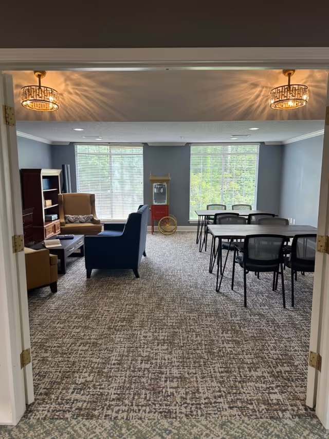 Interior view of a common area in an assisted living facility featuring a seating area with armchairs and a sofa on the left, a popcorn machine against the back wall, and two tables with chairs on the right side. The room has large windows with blinds letting in natural light, patterned carpet flooring, and two ceiling light fixtures.
