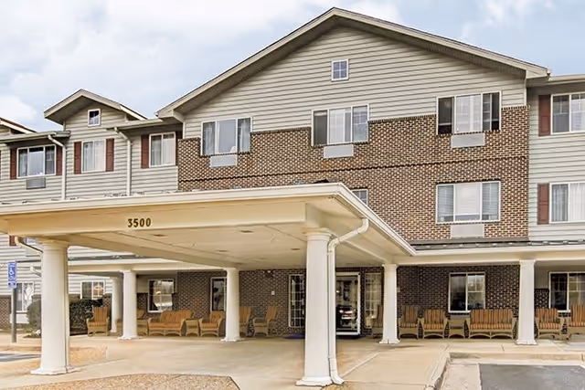 Front exterior view of a senior living facility building with a covered entrance supported by white columns. The building has a combination of brick and siding exterior with multiple windows and several wooden benches arranged under the covered area.