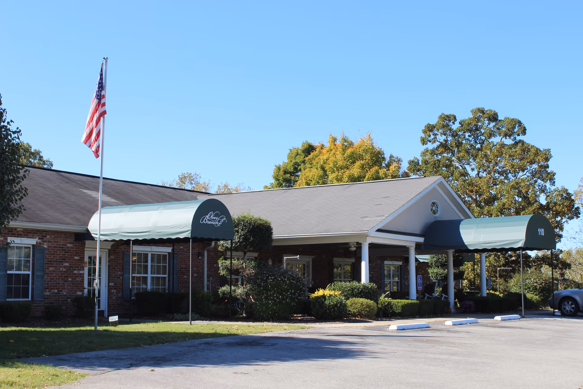 Front entrance of a one-story brick senior living building with green awnings, landscaping, and an American flag.