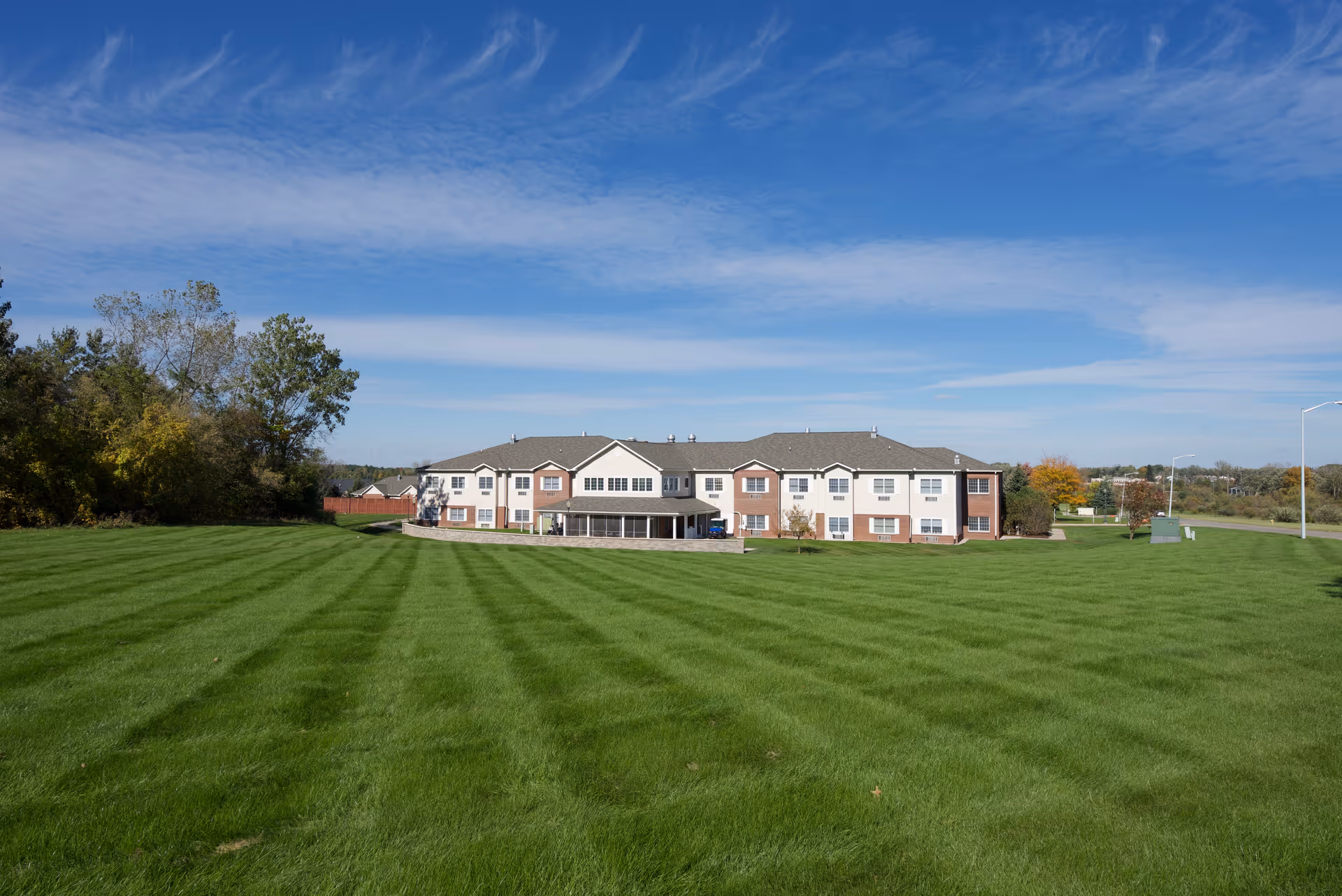 Wide shot of a two-story senior living building set behind a large striped lawn under a blue sky.