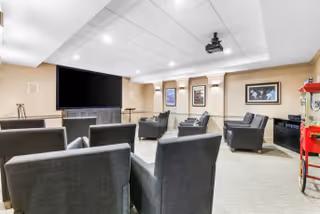 A cozy media room with multiple gray armchairs arranged facing a large flat-screen TV mounted on a wooden cabinet. The room has beige walls adorned with framed pictures, a ceiling-mounted projector, and a popcorn machine in the corner.