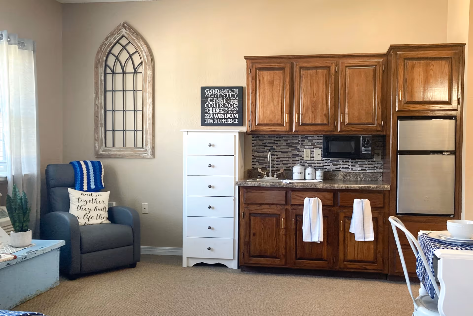 Cozy studio interior with a wooden kitchenette (sink, microwave, mini-fridge), a gray armchair, a white chest of drawers, and decorative wall accents.