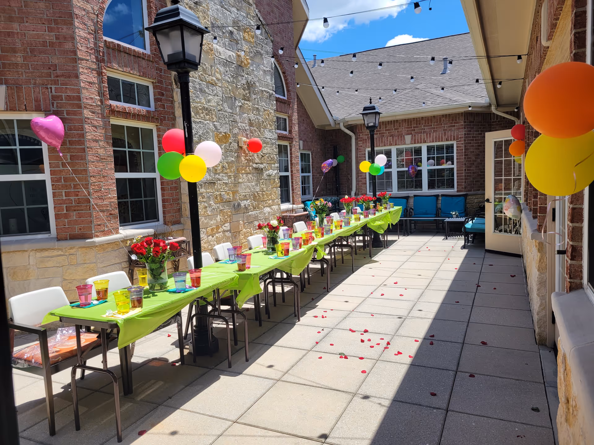 Outdoor patio area at Cypress Assisted Living - CyFair decorated for a celebration with a long table covered in a green tablecloth, colorful cups, flower vases, and balloons attached to chairs and walls. There are string lights overhead and rose petals scattered on the tiled floor.