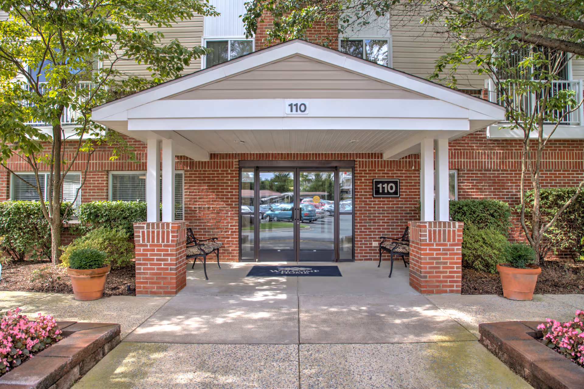Brick-front covered entrance numbered 110 with double glass doors, benches, and potted plants.