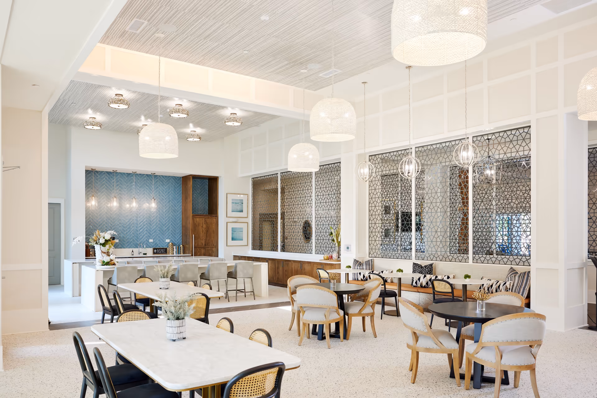 Bright and modern dining area with multiple tables and chairs, pendant lights hanging from the ceiling, a bar counter with stools, and decorative wall panels with geometric patterns.