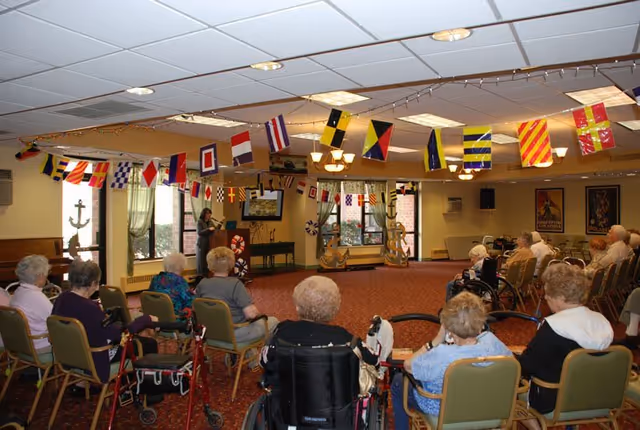 A group of elderly people seated in chairs and wheelchairs in a decorated room with nautical flags hanging from the ceiling. A woman stands at a podium near a window, addressing the group. The room has carpeted floors, large windows, and wall decorations including anchors and framed posters.