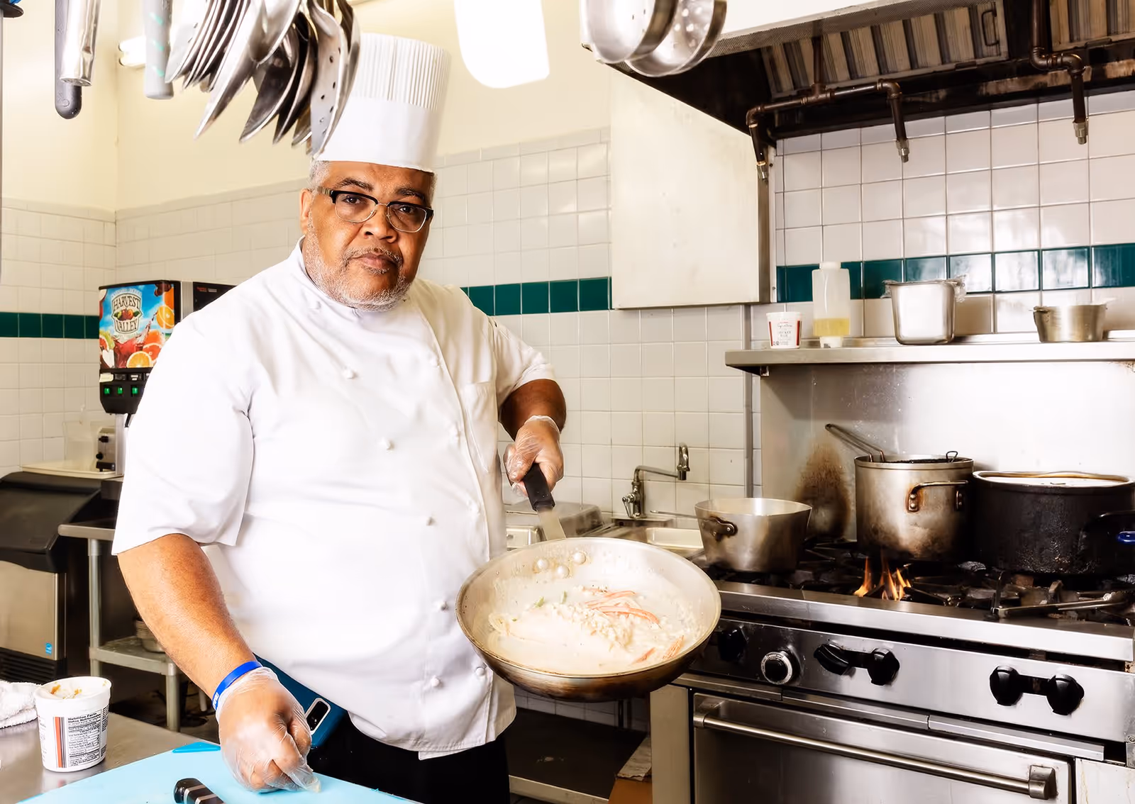 A chef wearing a white uniform and hat is cooking in a commercial kitchen, holding a frying pan with food over a stove with pots and pans.