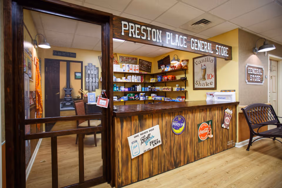 Interior view of Preston Place General Store, a small vintage-style store with wooden counter decorated with retro signs. Shelves behind the counter are stocked with various snacks and household items. The store has warm lighting and a cozy atmosphere with a bench nearby and a door with glass panels.