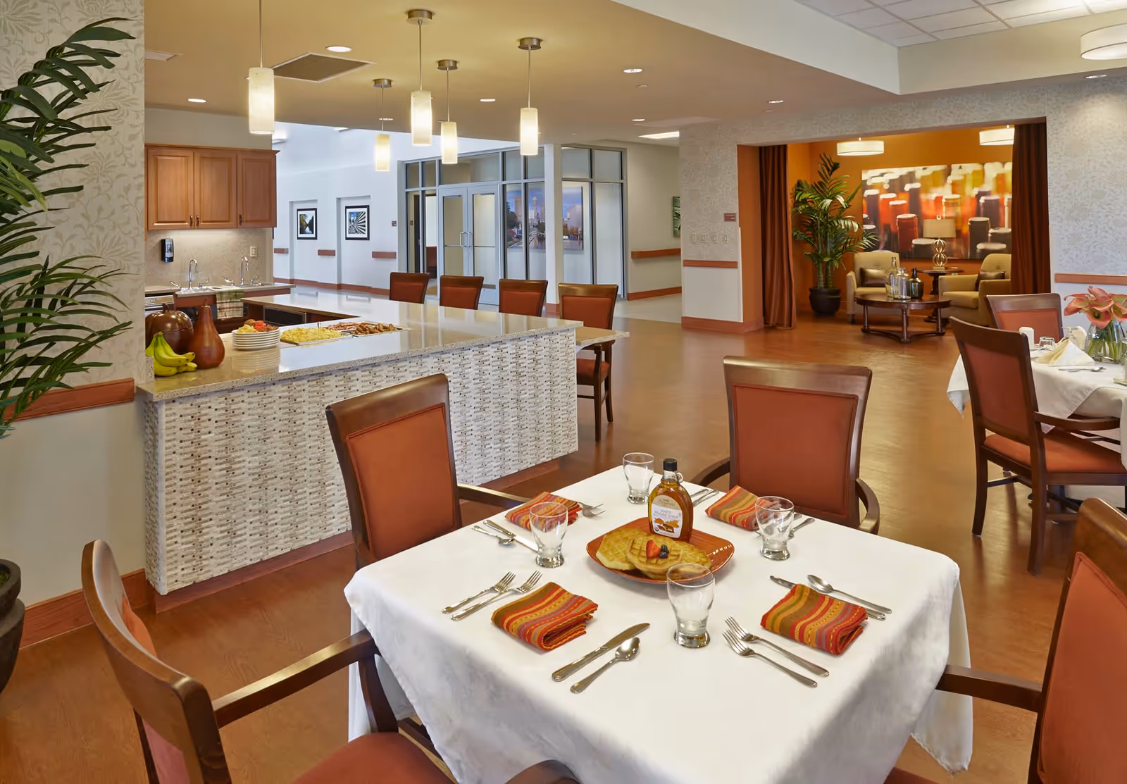 A dining area in Arlington Place Health Campus featuring a table set for four with plates, glasses, silverware, and striped napkins. The table has a plate of pancakes with syrup. In the background, there is a kitchen counter with fruit and snacks, several chairs, and a seating area with armchairs and a large colorful abstract painting on the wall. The room has warm lighting and wooden flooring.