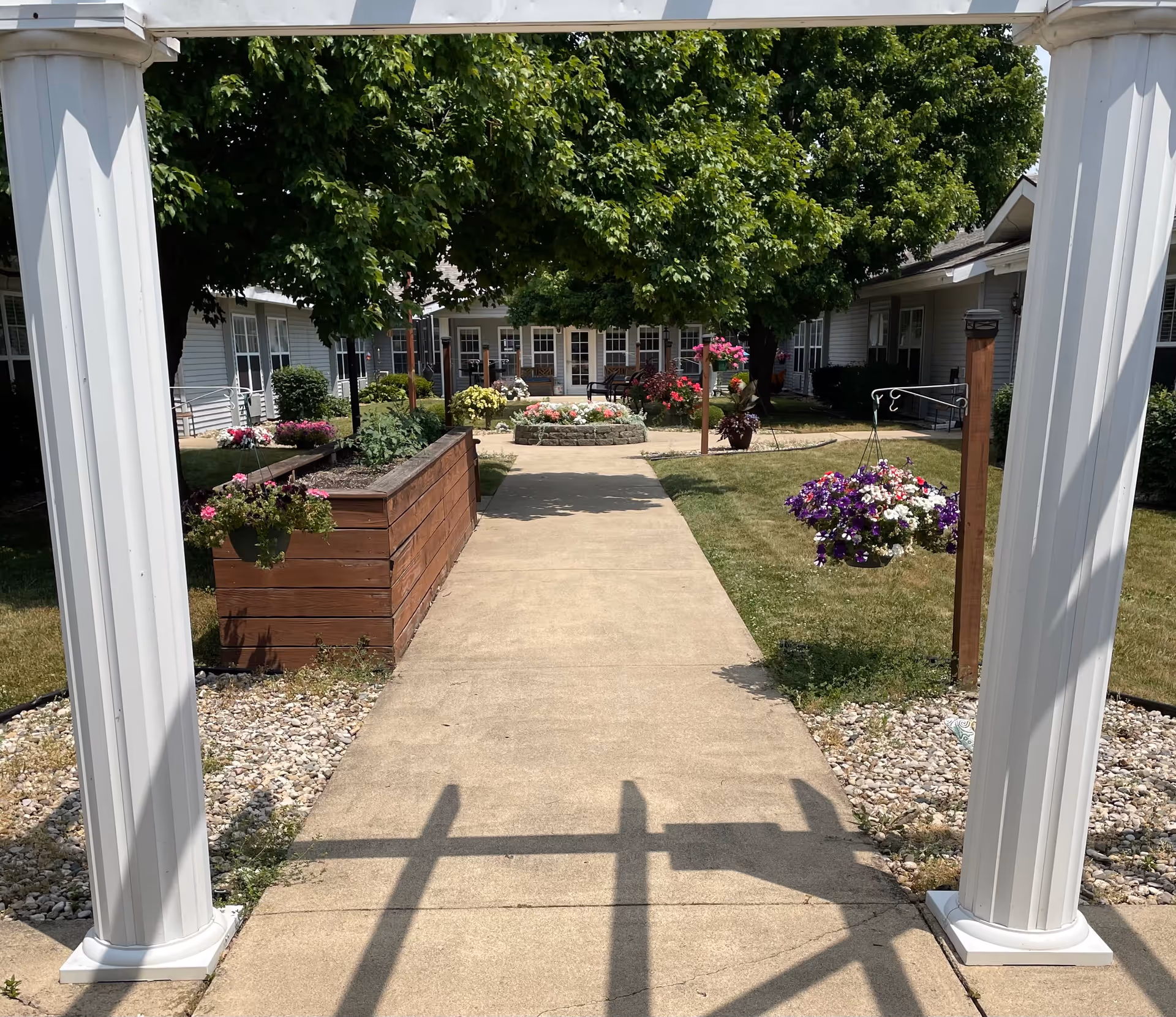 A sunny outdoor courtyard at Cedar Creek of Logansport featuring a concrete walkway flanked by flower beds and hanging flower baskets. There are green trees providing shade, and buildings with windows surround the courtyard.