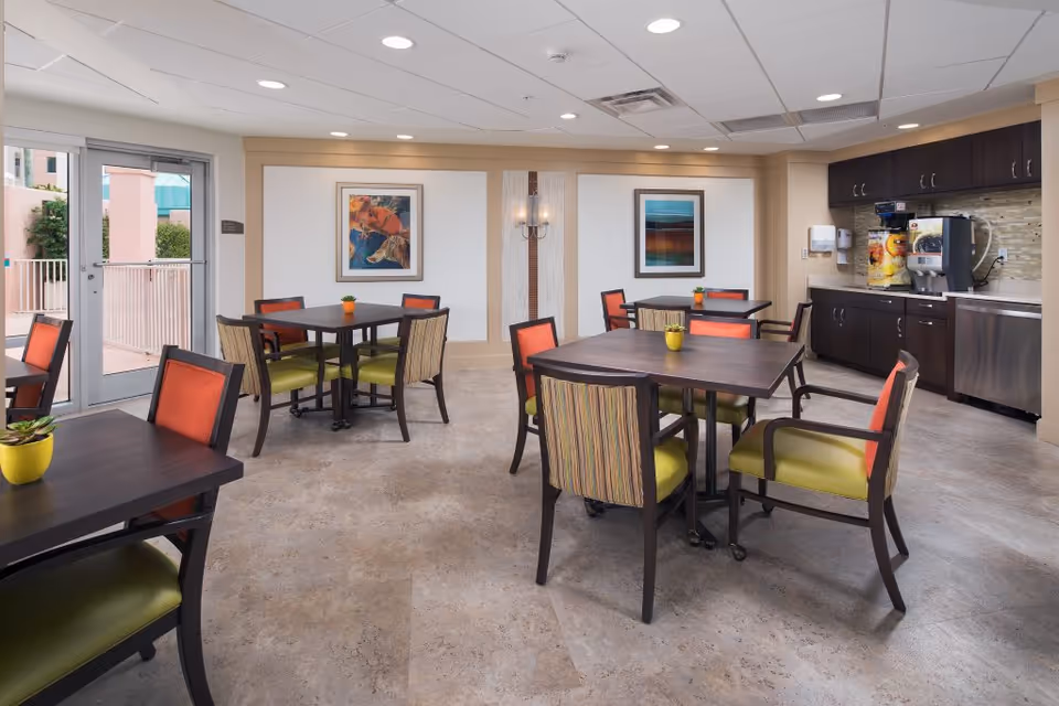Bright communal dining area with multiple square tables and colorful chairs near a kitchenette and beverage station.