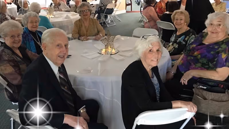 A group of elderly people sitting around a round table covered with a white tablecloth in a communal dining area. The table is set with napkins and a decorative centerpiece with candles. The seniors are smiling and appear to be enjoying a social gathering.