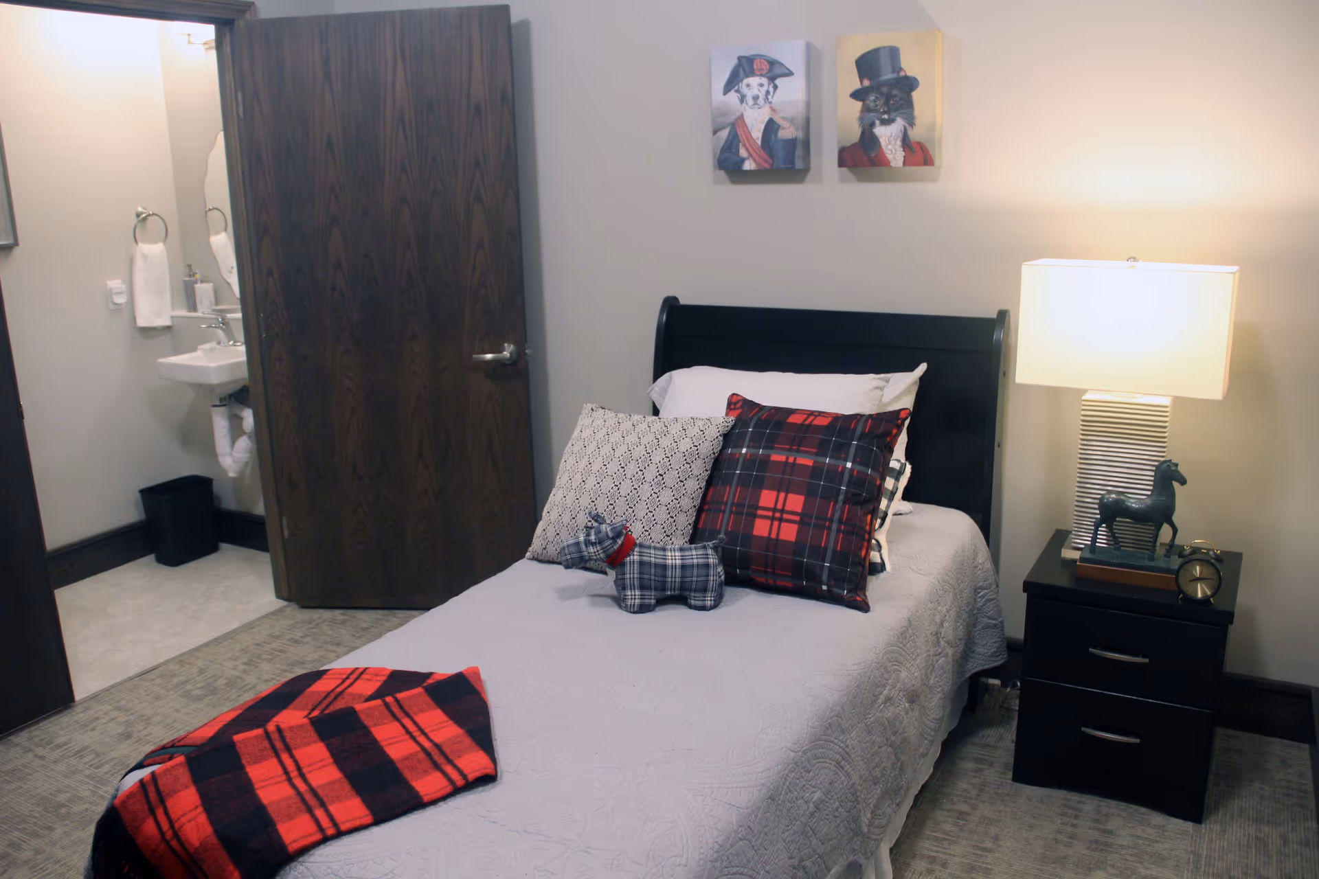 A cozy bedroom with a single bed covered in a light gray quilt and decorated with patterned pillows and a small plaid dog plush. A red and black plaid blanket is draped over the foot of the bed. Next to the bed is a black nightstand with a lamp, a small horse figurine, and a clock. The room has beige walls with two framed pictures of animals dressed in formal attire. An open door reveals a small bathroom with a sink, mirror, and towel ring.