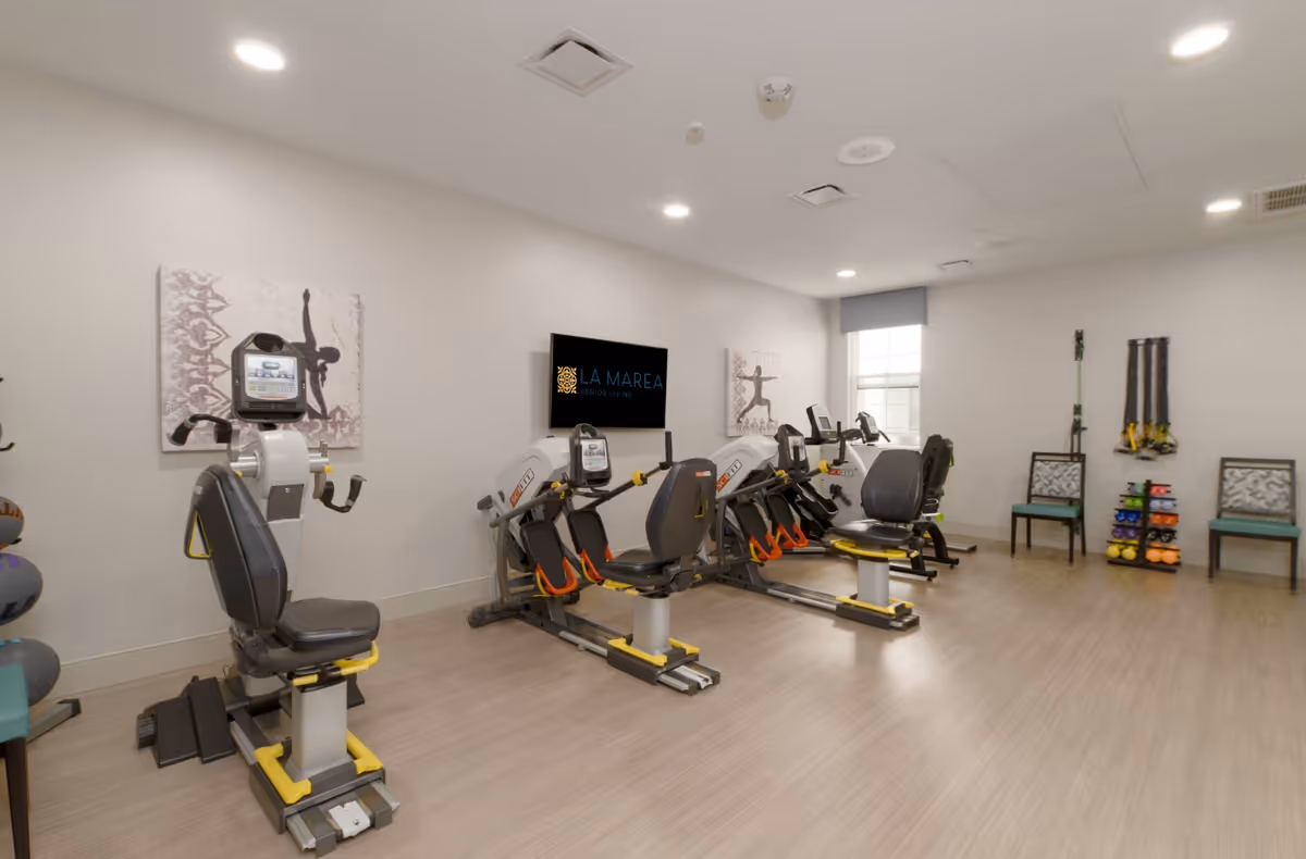 A bright fitness room at La Marea Senior Living featuring several exercise machines including recumbent bikes and rowing machines. The room has light-colored walls and flooring, a window with a blue valance, two chairs, and a rack with colorful dumbbells and resistance bands. A wall-mounted TV displays the La Marea Senior Living logo.