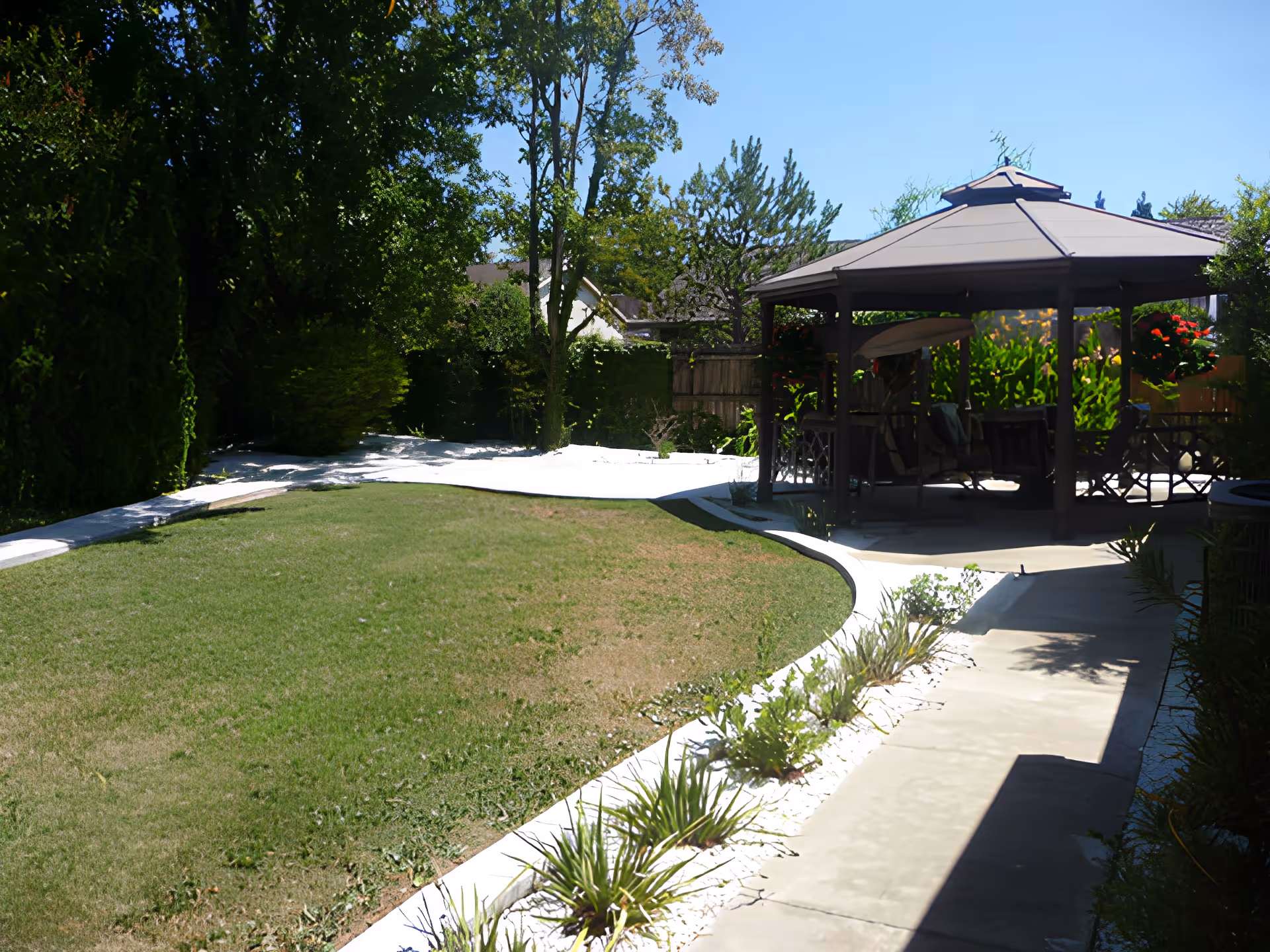 Outdoor garden area with a green lawn, a paved walkway, and a gazebo with seating underneath. The area is surrounded by trees and bushes under a clear blue sky.