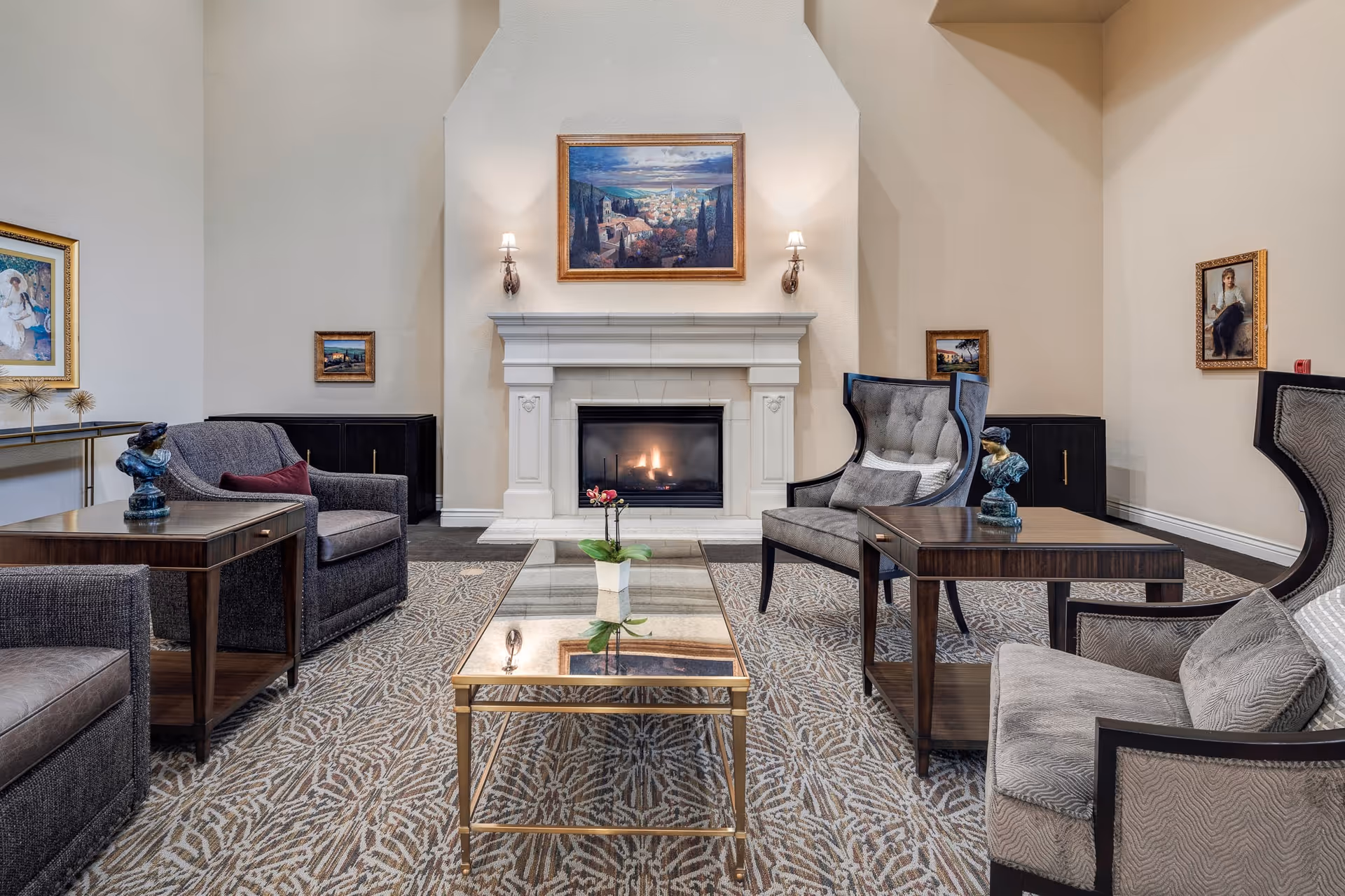 A cozy living room area with a lit fireplace centered on the back wall. The room features a patterned carpet, a glass coffee table with a small plant, two wooden side tables with decorative sculptures, and four upholstered chairs arranged around the tables. The walls are adorned with framed paintings and the room has a warm, inviting atmosphere.