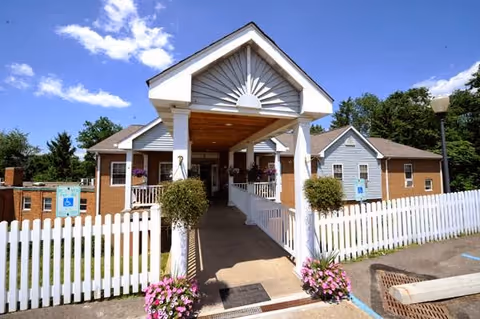Front exterior view of Norbert Personal Care facility showing a covered entrance with white columns and a white picket fence. There are flower pots with pink flowers on either side of the entrance ramp, a clear blue sky with some clouds, and a handicap parking sign visible near the building.