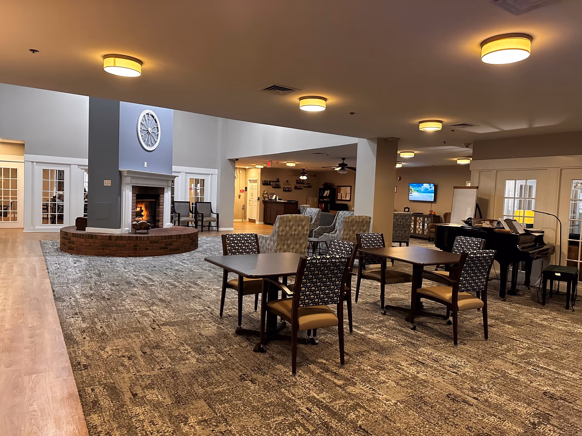A spacious common area in a senior living facility featuring a central brick fireplace with a large clock above it, several tables with chairs, comfortable armchairs, a piano, and a television mounted on the wall. The room has warm lighting and a carpeted floor with wood flooring around the edges.