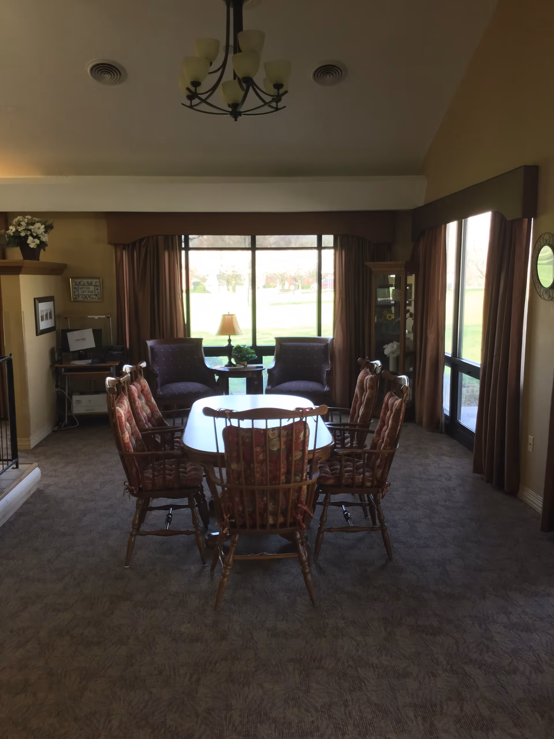 A communal room with a wooden dining table surrounded by upholstered chairs, two armchairs by a large window, and a chandelier overhead.