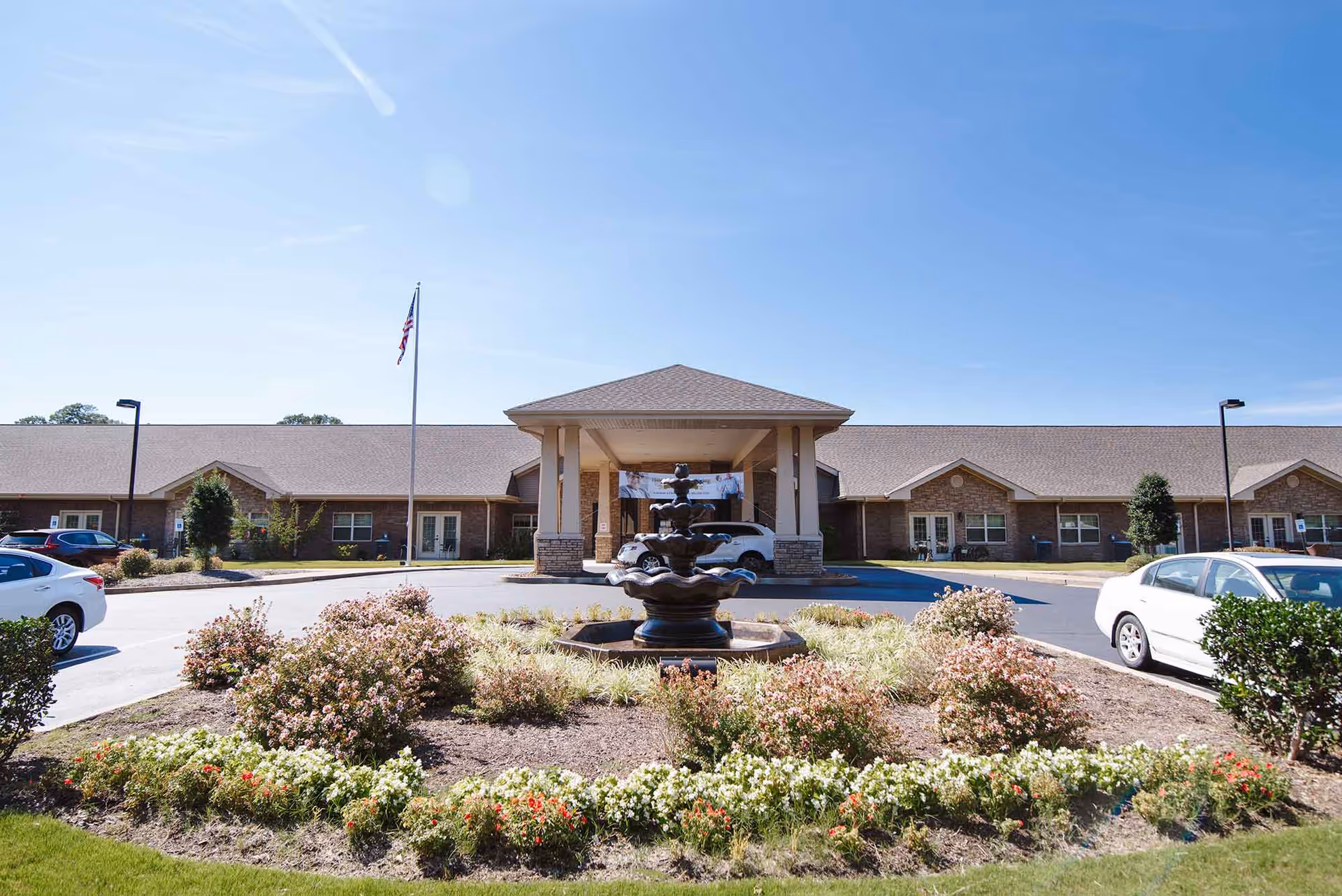 Front exterior view of Hearthside Senior Living facility with a central covered entrance, a flagpole with an American flag, a decorative fountain surrounded by landscaped flowers and bushes, and parked cars on either side of the driveway.