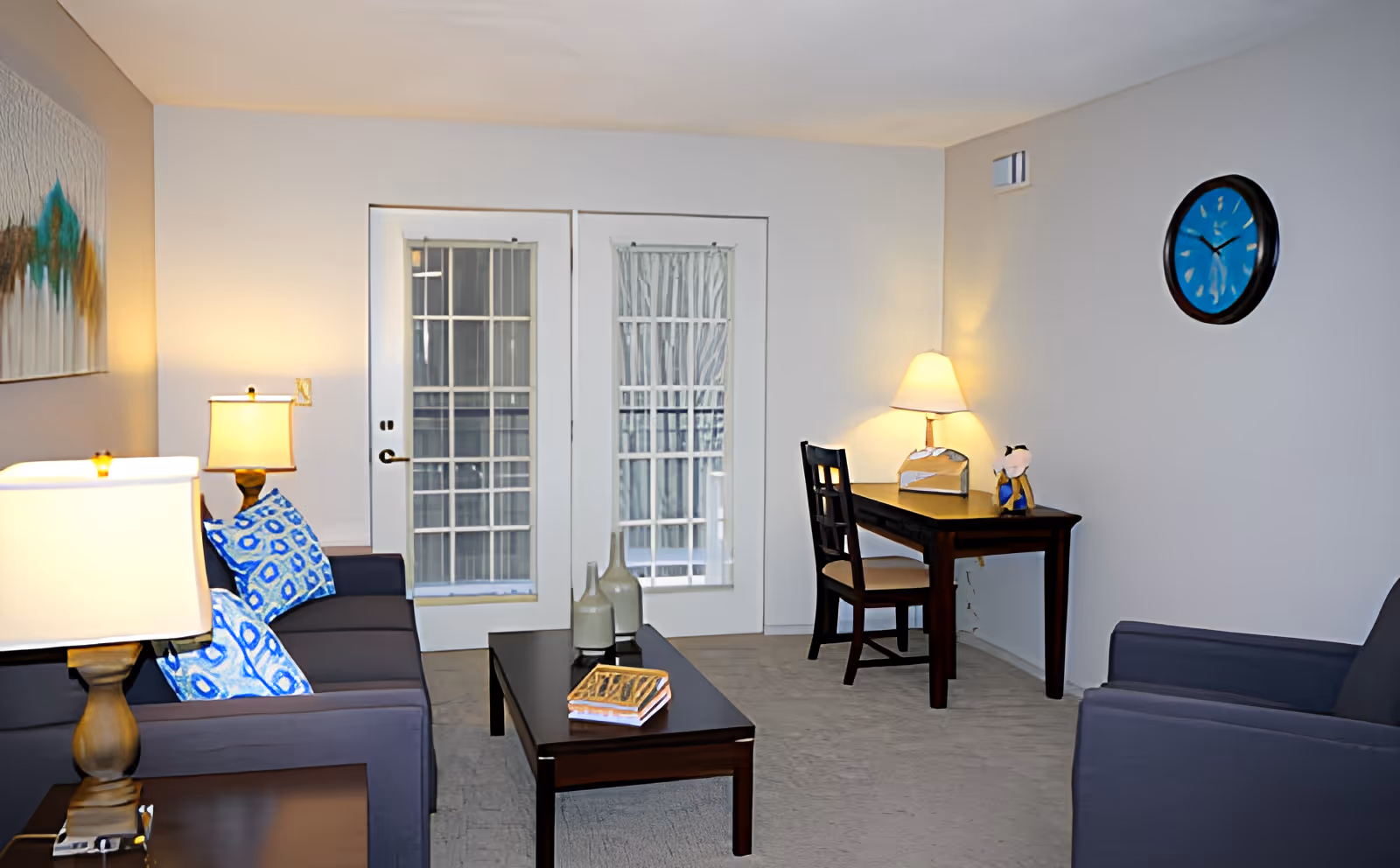 A cozy living room with two dark blue sofas adorned with blue patterned pillows, a wooden coffee table with decorative vases and a book, a wooden desk with a chair and a lamp, a wall clock with a blue face, and French doors with glass panes leading outside.