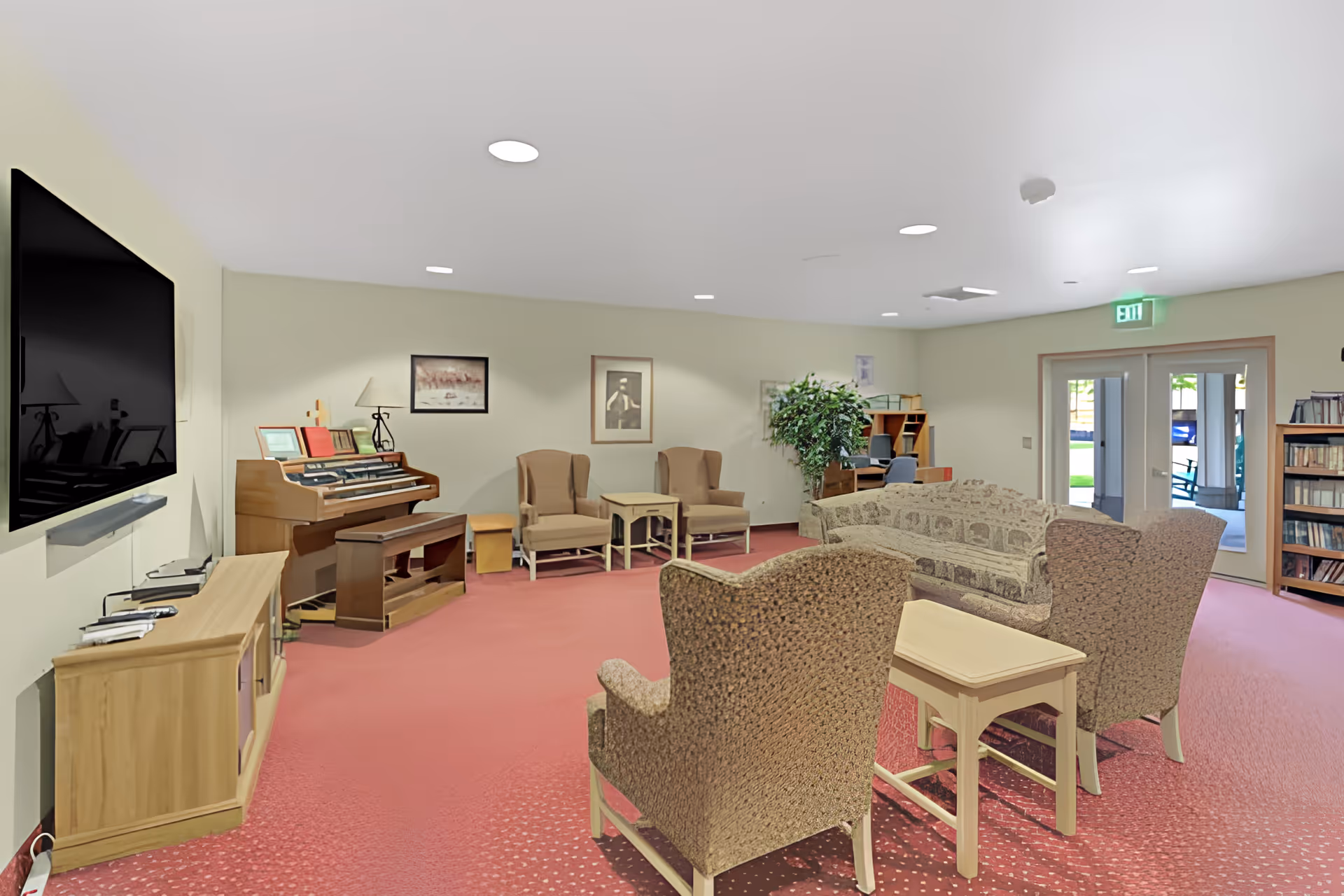 A spacious living room area with a red carpet, beige walls, and recessed ceiling lights. The room features a large flat-screen TV mounted on the wall above a wooden cabinet, a wooden piano with a bench, several armchairs and sofas arranged around small tables, a bookshelf filled with books, and a potted plant. Double glass doors lead outside to a patio area with rocking chairs.