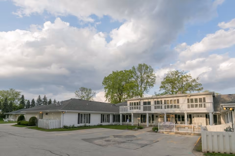 White two-story assisted living building with balconies, a fenced patio and paved driveway under a cloudy sky.