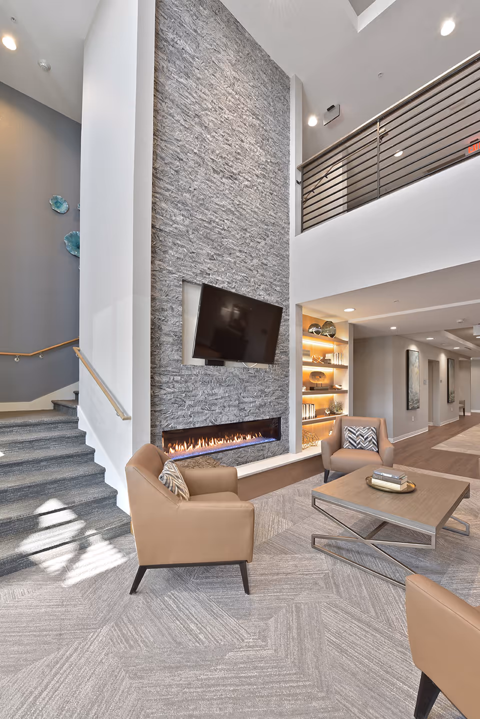 Modern senior living facility common area with a tall stone fireplace featuring a mounted flat-screen TV above a linear gas fireplace. The room includes beige armchairs with patterned cushions, a wooden coffee table with books, a carpeted floor, and a staircase with a wooden handrail. There is a mezzanine level with a metal railing and built-in shelves with decorative items and warm lighting.