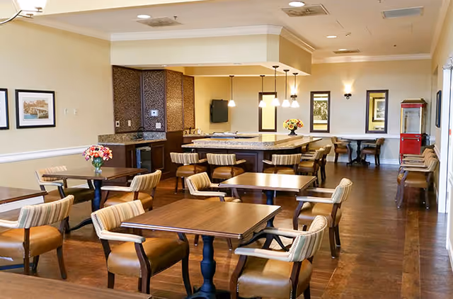 Communal dining room with several wood tables and upholstered chairs, a central serving island with pendant lights and wall decor.