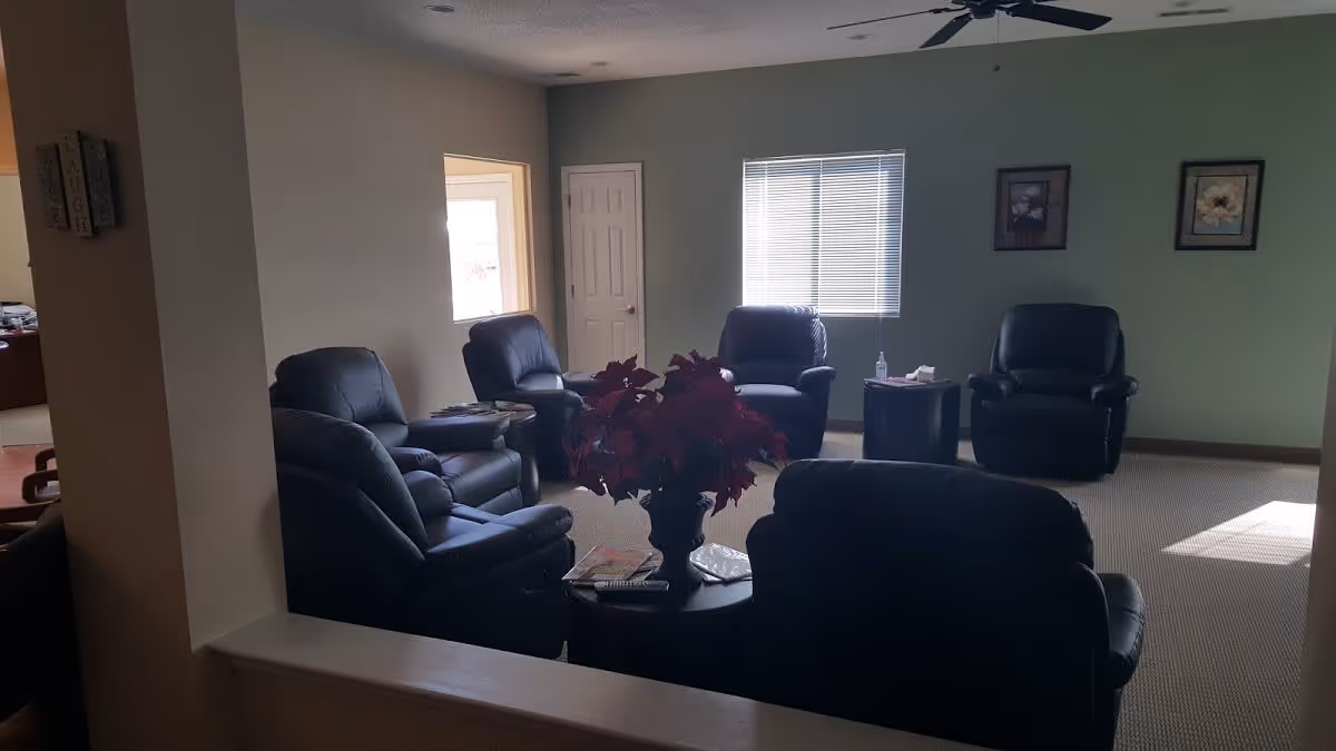 A quiet living room area with six black leather recliners arranged in a circle around two small round tables. One table has a vase with red flowers and some magazines, while the other has a bottle of hand sanitizer and tissues. The room has beige walls, a window with blinds, two framed floral pictures on the wall, and a ceiling fan.
