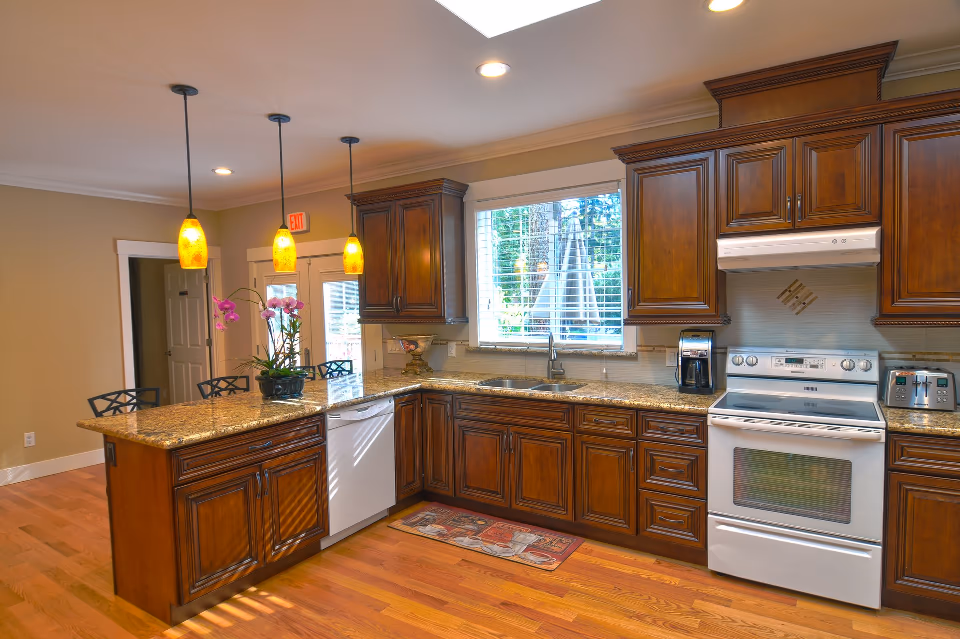 A spacious kitchen with wooden cabinets, granite countertops, and hardwood floors. The kitchen features a white stove, dishwasher, toaster, coffee maker, and a window with blinds above the sink. Three pendant lights hang over a kitchen island with bar stools and a potted orchid plant on top.