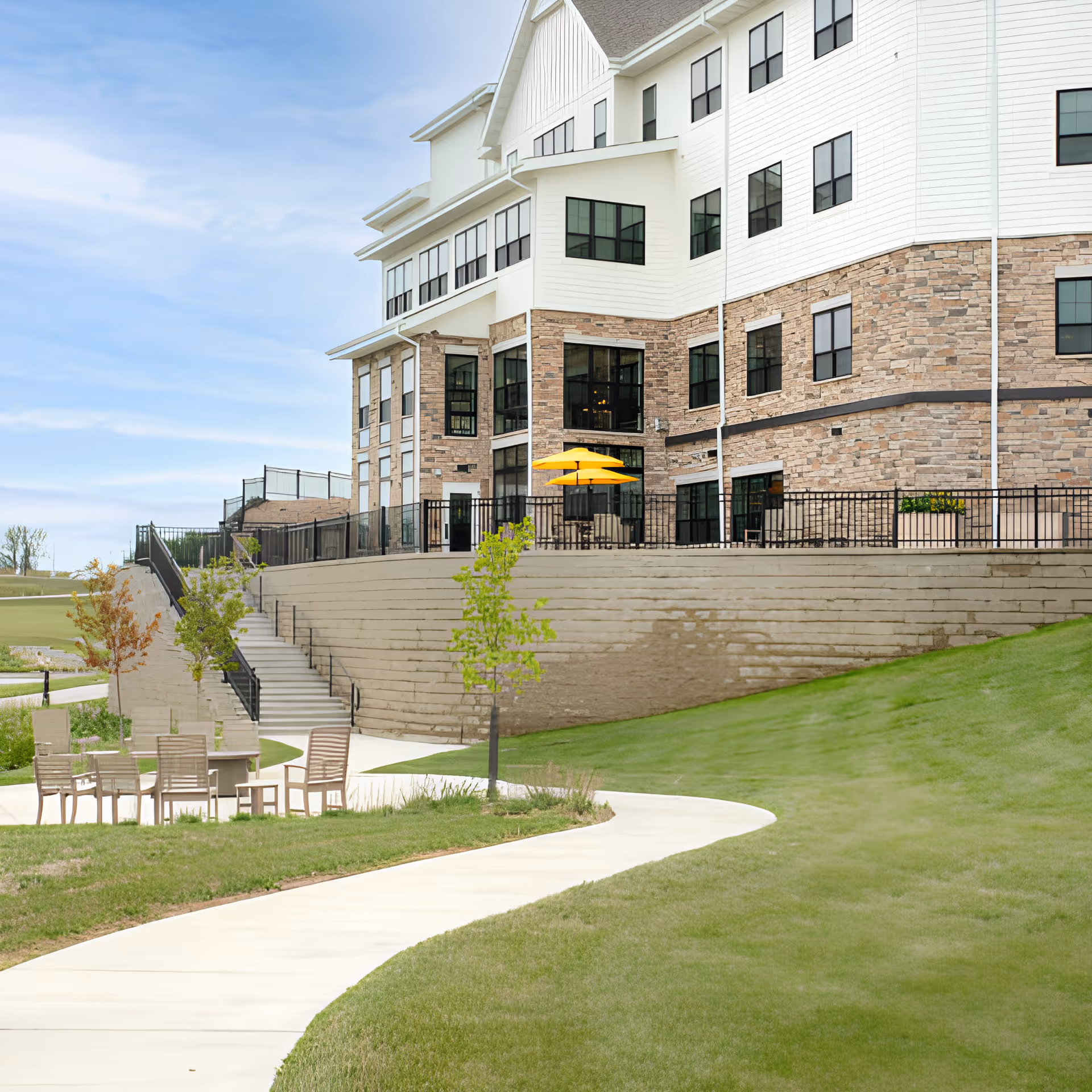 Exterior view of a senior living facility building with a stone and white siding facade, multiple windows, and a patio area with yellow umbrellas. In the foreground, there is a curved concrete pathway, green grass, small trees, and outdoor seating with chairs and a table.