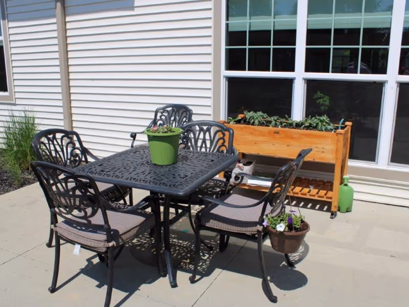 Outdoor patio area with a black metal table and four matching chairs with cushions. A green potted plant is on the table, and a wooden planter box with plants is placed against the building's white siding near large windows.